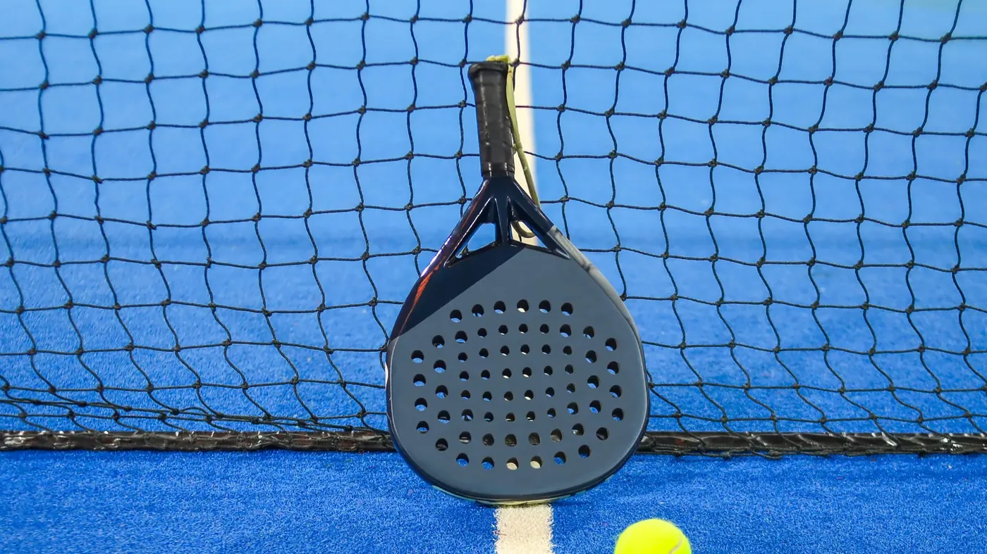 Pala de pádel apoyada en la red de una pista azul, con el grip de la empuñadura a la vista